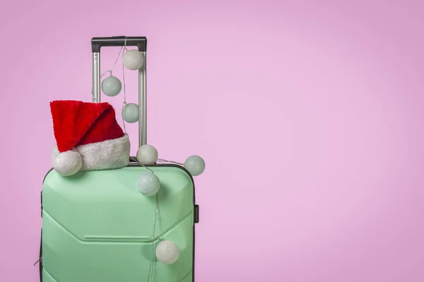 Walizka z tworzywa sztucznego, czapka Świętego Mikołaja i łańcuch na różowym tle. Koncepcja podróży, podróże służbowe, wycieczki do odwiedzenia przyjaciół i krewnych na święta Bożego Narodzenia. Podróż Sylwestrowa — Zdjęcie stockowe