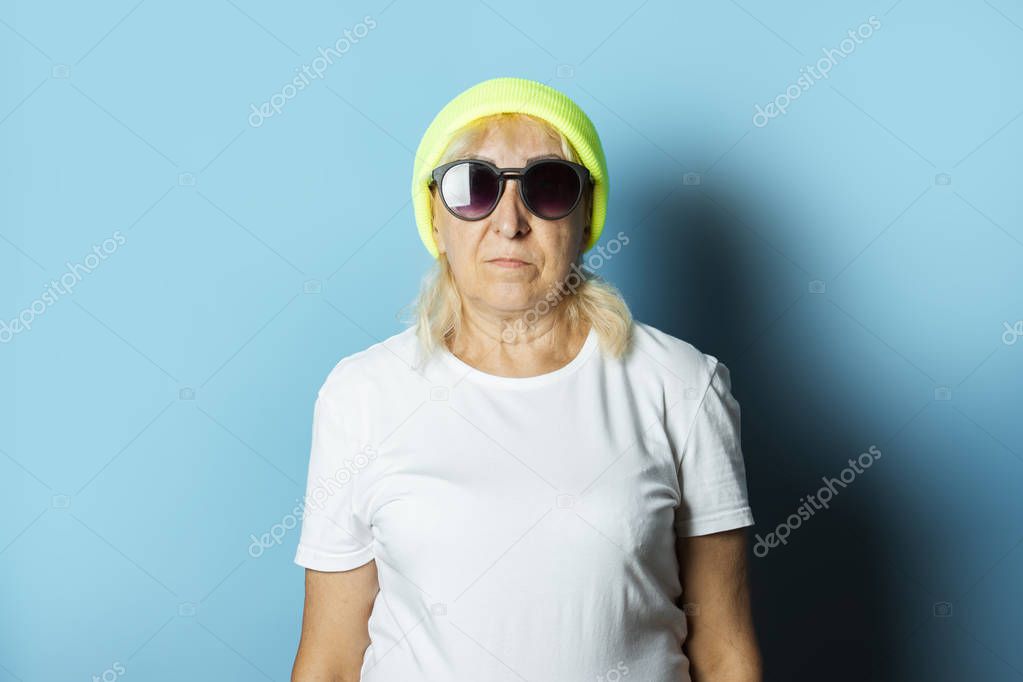 Funny old woman in a hat and glasses, makes a hand gesture on a blue background. Concept cool stylish grandmother, modern style