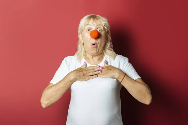 Rolig gammal kvinna med en röd näsa av en clown på en rosa bakgrund. Concept röd näsa dag, semester, fest, clown — Stockfoto