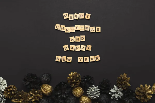 Cônes de pin et noix peints dans des couleurs dorées, noires, blanches sur un fond noir. Ajout du texte Bonne année et Joyeux Noël. Couché plat, vue du dessus — Photo