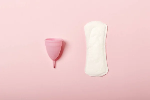 Cangkir menstruasi dan pembalut wanita dengan latar belakang merah muda. Konsep menstruasi, pilihan antara produk kebersihan feminin. Letak datar, atas — Stok Foto