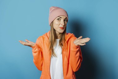 Kapüşonlu ve şapkalı güzel bir genç kadın ellerini mavi arka planda gezdirerek bir jest yapıyor. Kış konsepti, Noel, kış tatili, soğuk hava başlangıcı.