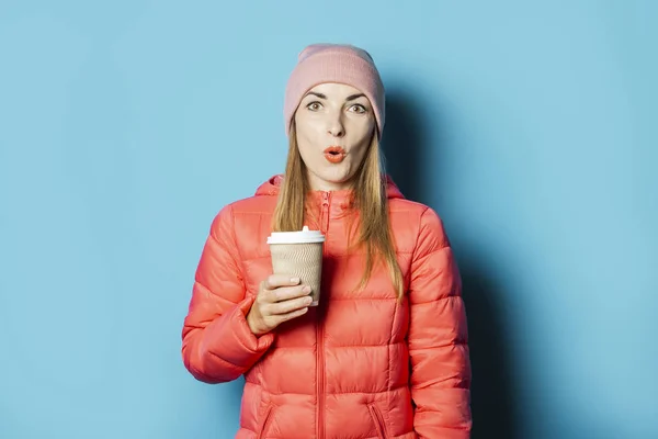 Freezing Beautiful Young woman in a hat and a winter jacket holds a cup with hot coffee on a blue background. Winter concept, season change, hot drinks