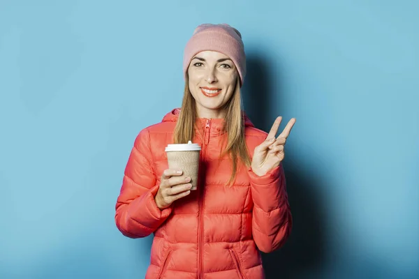 모자와 겨울 재킷을 입은 아름다운 청녀가 파란색 배경에 뜨거운 커피가 있는 컵을 들고 있다. 겨울 컨셉, 계절 변경, 따뜻한 음료 — 스톡 사진