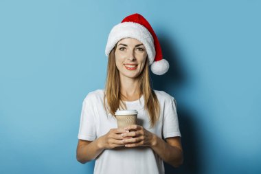 Noel Baba şapkası bir gülümseme ile Güzel Genç kadın mavi bir arka plan üzerinde kahve bir fincan tutar. Yeni Yıl ve Noel için Konsept fikir, Noel hediyeleri