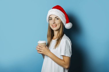 Noel Baba şapkası bir gülümseme ile Güzel Genç kadın mavi bir arka plan üzerinde kahve bir fincan tutar. Yeni Yıl ve Noel için Konsept fikir, Noel hediyeleri