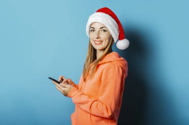 Kapüşonlu ve Noel Baba şapkalı güzel bir genç kadın mavi arka planda bir cep telefonu tutuyor. Kış kavramı, Noel, kış tatili, başvuru