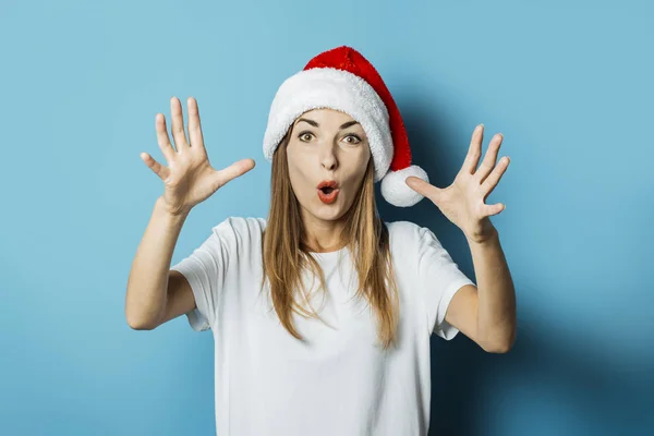 Hermosa mujer joven con una cara sorprendida en un sombrero de Santa Claus sobre un fondo azul. Concepto de idea para el año nuevo y la Navidad — Foto de Stock