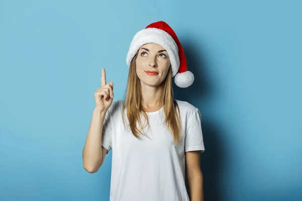 Mujer joven en sombrero de Santa Claus con una cara misteriosa sobre un fondo azul. Concepto de idea para el año nuevo y la Navidad — Foto de Stock