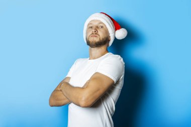 Mavi arka planda kollarını kavuşturmuş Noel Baba şapkalı bir adam..