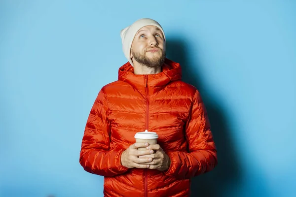 Man with a pensive face in a hat and a red jacket holds a cup of