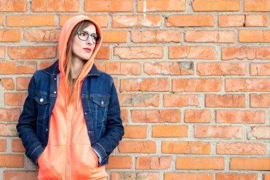 Gözlüklü, sweatshirt ve kot ceket giymiş, elleri cebinde bir sweatshirt tutan, kırmızı tuğlalı duvara bakan ciddi bir genç kadın..