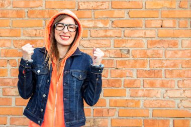 Gözlüklü neşeli genç bir kadın, kapalı gözlü yumruklar tutuyor, kırmızı tuğladan bir duvarda sweatshirt ve kot ceket giyiyor..