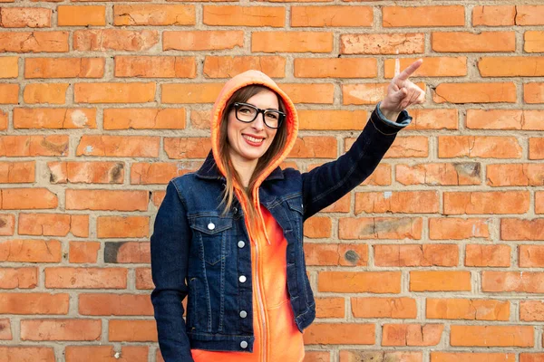Happy young girl with glasses points a finger to the side, wearing a sweatshirt and denim jacket on a background of a red brick wall.
