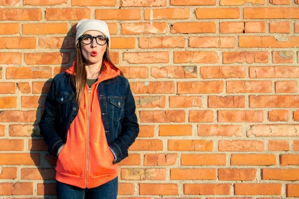Shocked young girl in glasses, a light hat and a denim jacket, holds her hands in a sweatshirt and lay her back against a red brick wall.