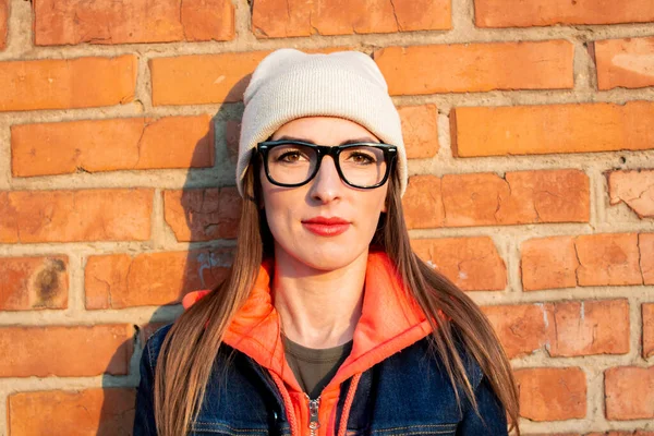 Young woman smiling with glasses against a brick wall, wearing a gray hat, a sweatshirt and a denim jacket, close-up.