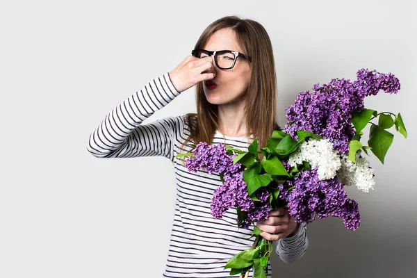 Young woman holds a lilac pinched her nose with fingers, allergic to flowers, on a light background