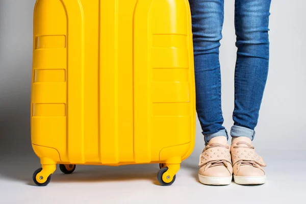 Female legs in jeans stand next to a yellow suitcase on a light background. Travel concept, flight expectation, vacation. Only legs are visible. Banner.