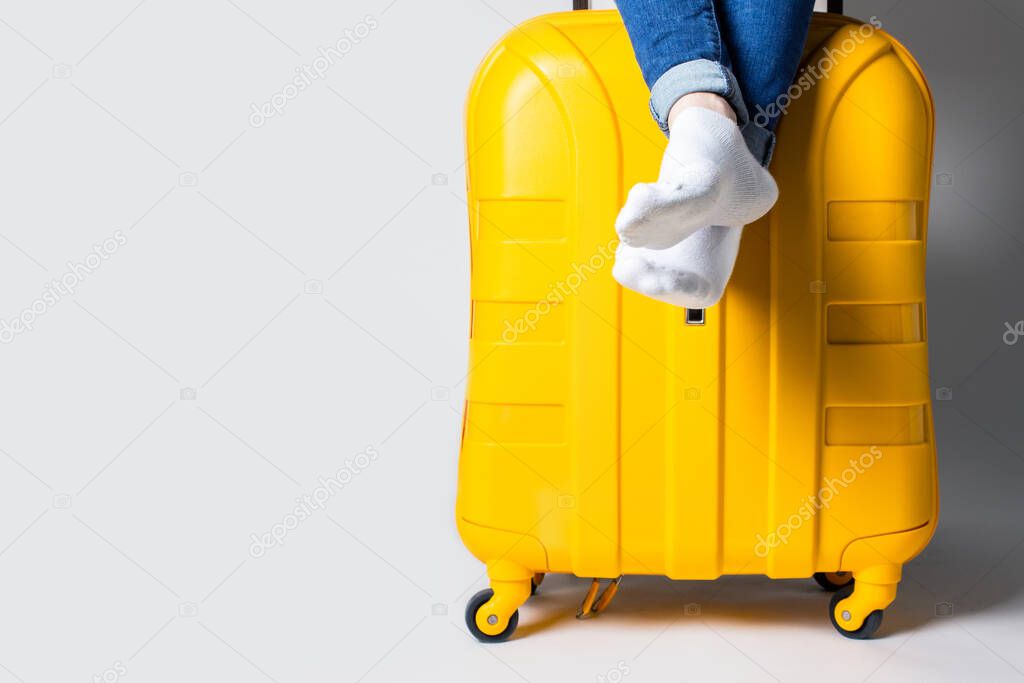 Woman is sitting on a yellow plastic suitcase on a light background. Travel concept, flight expectation, vacation. Only legs are visible. Banner.