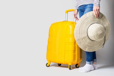 Kadının elinde plastik sarı bir bavul ve arka planda güneşten kalma bir şapka var. Seyahat konsepti, uçuş beklentisi, tatil. Sadece bacaklar görünür. Pankart.