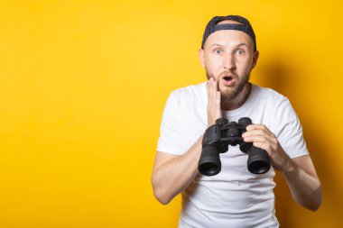 Beyaz tişörtlü ve beysbol şapkalı genç adam, şaşırmış ve şok olmuştu. Sarı arka planda dürbün tutuyordu.