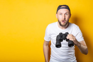 Beyaz tişörtlü ve beysbol şapkalı genç adam, şaşırmış ve şok olmuştu. Sarı arka planda dürbün tutuyordu.