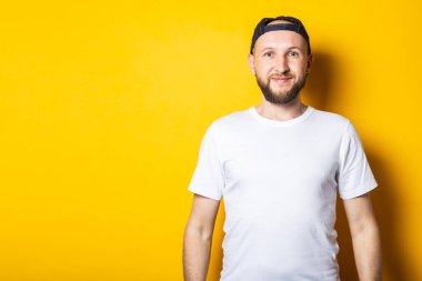 Genç adam, beyaz tişörtlü ve sarı arka planda duran beyzbol şapkalı arkadaş canlısı ve mutlu..