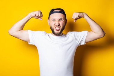 Genç adam beyaz bir tişört giyiyor, güçlü, atletik, sarı bir arka planda pazısı var. Gücün göstergesi, atletik yapı.