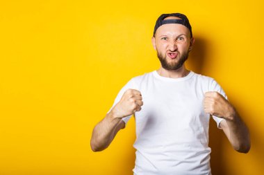 Beyaz tişörtlü genç adam, güçlü, atletik, sarı arka planda yumruklarını gösteriyor. Gücün göstergesi, atletik yapı.