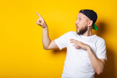 Beyaz tişörtlü ve beysbol şapkalı genç adam, sarı arka planda bir parmak gösteriyor..