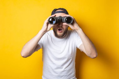 Beyaz tişörtlü, şoktan zevk alan genç adam sarı arka planda dürbünle şaşırmış görünüyor..
