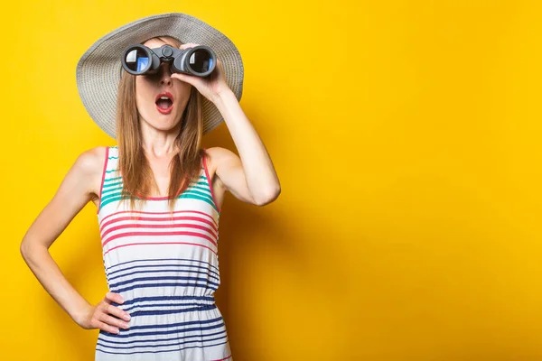 Young woman in shock in surprise with a hat and a striped dress looks in surprise with binoculars on a yellow background.