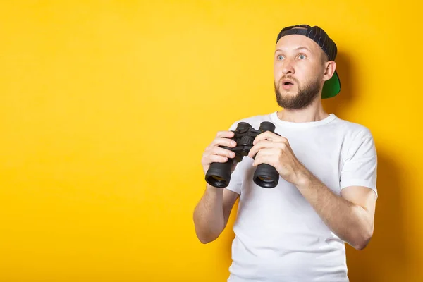 Young man in a white T-shirt and a baseball cap, surprised looks away holds binoculars on a yellow background