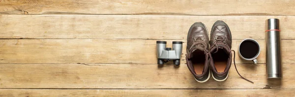 Boots for trail, binoculars, thermos, cup with tea on a wooden background. The concept of hiking, tourism, camp, mountains, forest. Banner. Flat lay, top view.