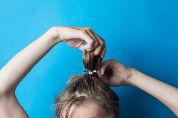 Girl straightens the disheveled tail of hair on her head, against a blue background. Modern fast hairstyle. Hair tied with elastic band.