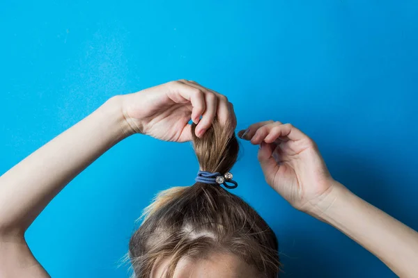 Girl straightens the disheveled tail of hair on her head, against a blue background. Modern fast hairstyle. Hair tied with elastic band.