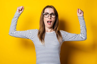 Yumruklarını sıkmış ve kollarını kaldırmış genç bir kadın sarı arka planda seviniyor..