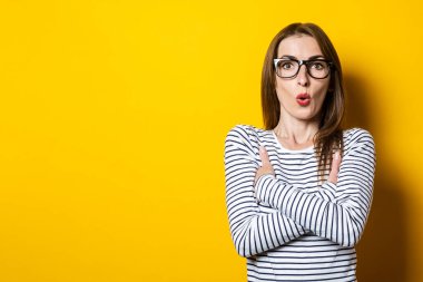 Şokta olan genç bir kadın sarı arka planda kollarını kavuşturuyor..