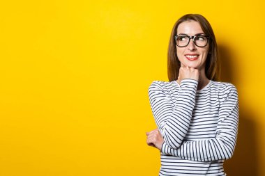 Dost canlısı genç bir kadın çenesine yumruk atarak gülümsüyor ve sarı bir arka plana bakıyor..