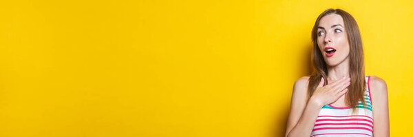Surprised young woman in a striped dress holds palms on the heart on a yellow background. Banner.