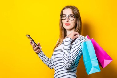 Dost canlısı genç bir kadın sarı arka planda alışveriş çantaları ve telefon tutuyor.