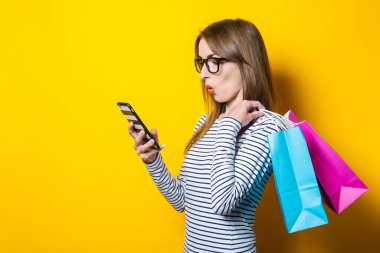Şokta olan genç bir kadın telefona şokta bakıyor, sarı arka planda paketleri elinde tutuyor.