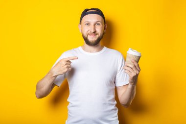 Dost canlısı gülümseyen genç adam sarı arka planda kahve ile kağıt bardağı işaret ediyor.