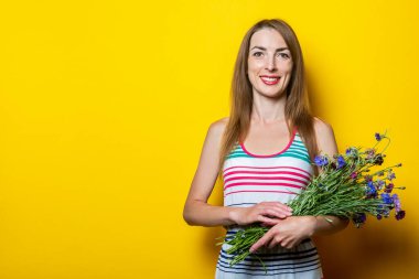 Sarı arka planda bir buket kır çiçeğiyle gülümseyen cana yakın genç kız.