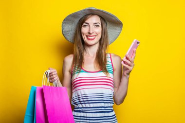 Sarı arka planda, elinde telefon ve paketler olan, şapkalı, mutlu, genç bir kadın.