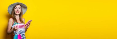 Sarı arka planda, elinde telefon ve paketler olan, şapkalı, mutlu, genç bir kadın. Pankart