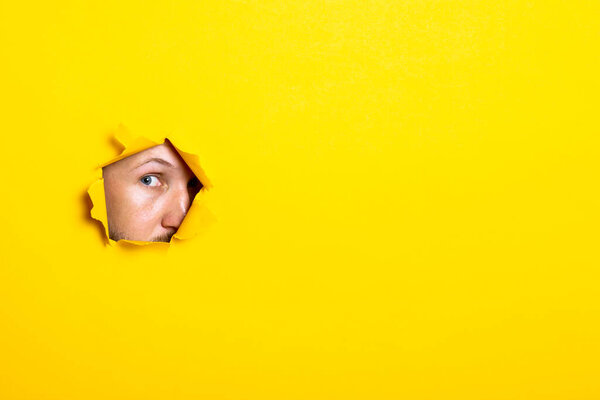 Young man looking with an eye into the hole of torn yellow background