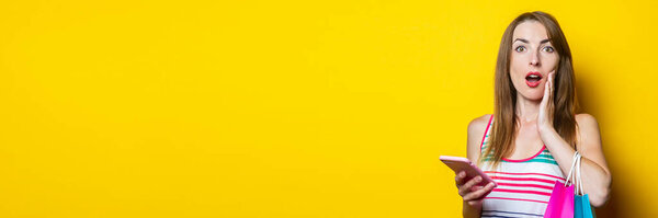 Surprised young woman holding hand on cheek with phone and shopping bags on yellow background. Banner