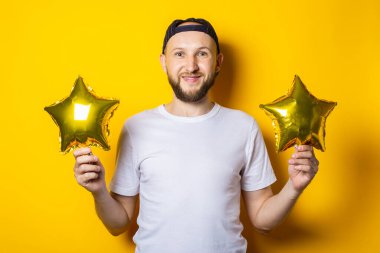 Sırıtkan sakallı genç adam sarı arka planda bir yıldız şeklinde balonlar tutuyor.