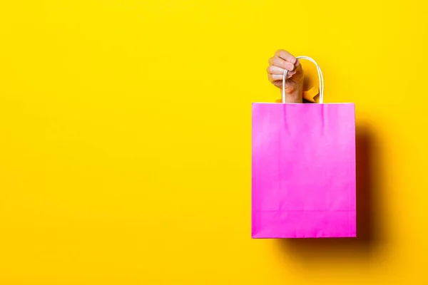 Female hand holds a pink package with purchases on a yellow background.